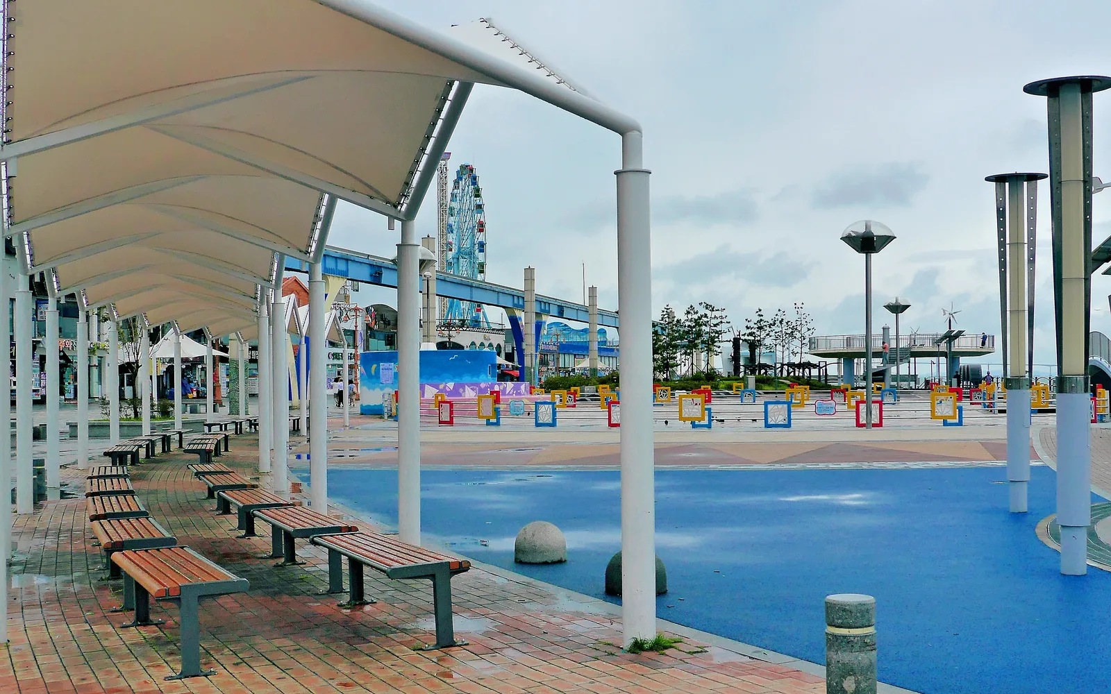 Covered benches and an open waterfront plaza on Wolmido with an amusement wheel and elevated track in the background